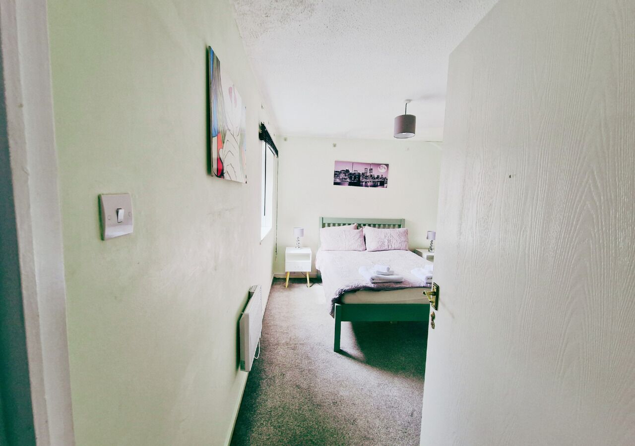 Ein schmaler Flur führt zu einem Bett mit Kopfteil, einem Nachttisch und einem an der Wand hängenden Bild.