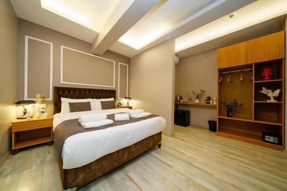 A modern hotel room with a large bed, wooden headboard, bedside tables with lamps, a built-in wardrobe, and a small desk with a lamp.