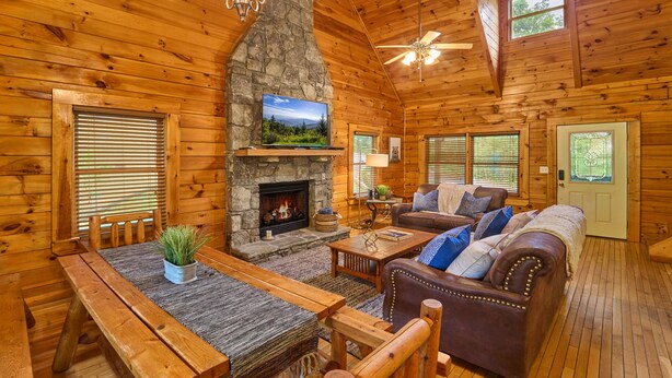 A cozy log cabin interior with a stone fireplace, wooden furniture, and a ceiling fan. - Mountain Escape Book Now (Pigeon Forge)