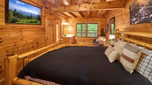 A wooden cabin interior with a large bed, a TV mounted on the wall, and a window with a view of greenery. - Mountain Escape Book Now (Pigeon Forge)
