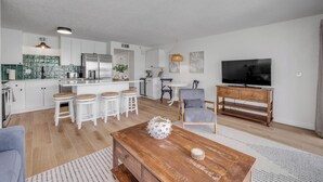 A modern kitchen with white cabinets, a central island, and a dining area with white stools. There is a wooden coffee table with decorative items and a TV on a wooden stand.