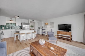 Une cuisine moderne avec des meubles blancs, un îlot central et un coin repas agrémenté de tabourets blancs. On y trouve une table basse en bois ornée d’objets décoratifs et un téléviseur posé sur un meuble en bois.