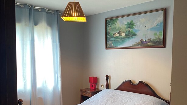 A bedroom with a wooden bed, a painting on the wall, a ceiling light, and a nightstand with a red object. - Marjolaine Bed and Breakfast on the ground floor (Lourdes)