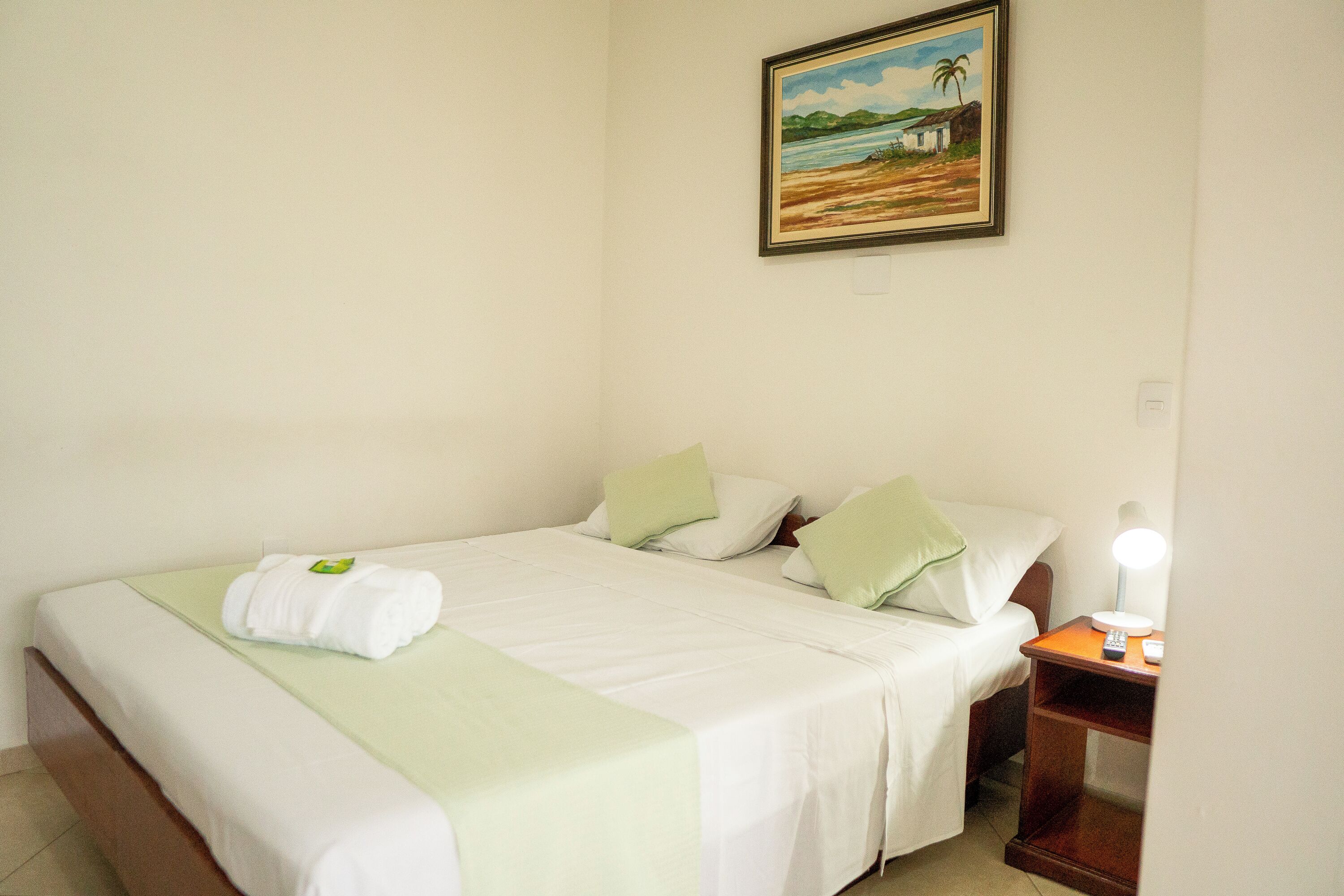 Una cama bien hecha con una colcha verde y sábanas blancas, una mesita de noche con una lámpara, y un cuadro de una playa en la pared.