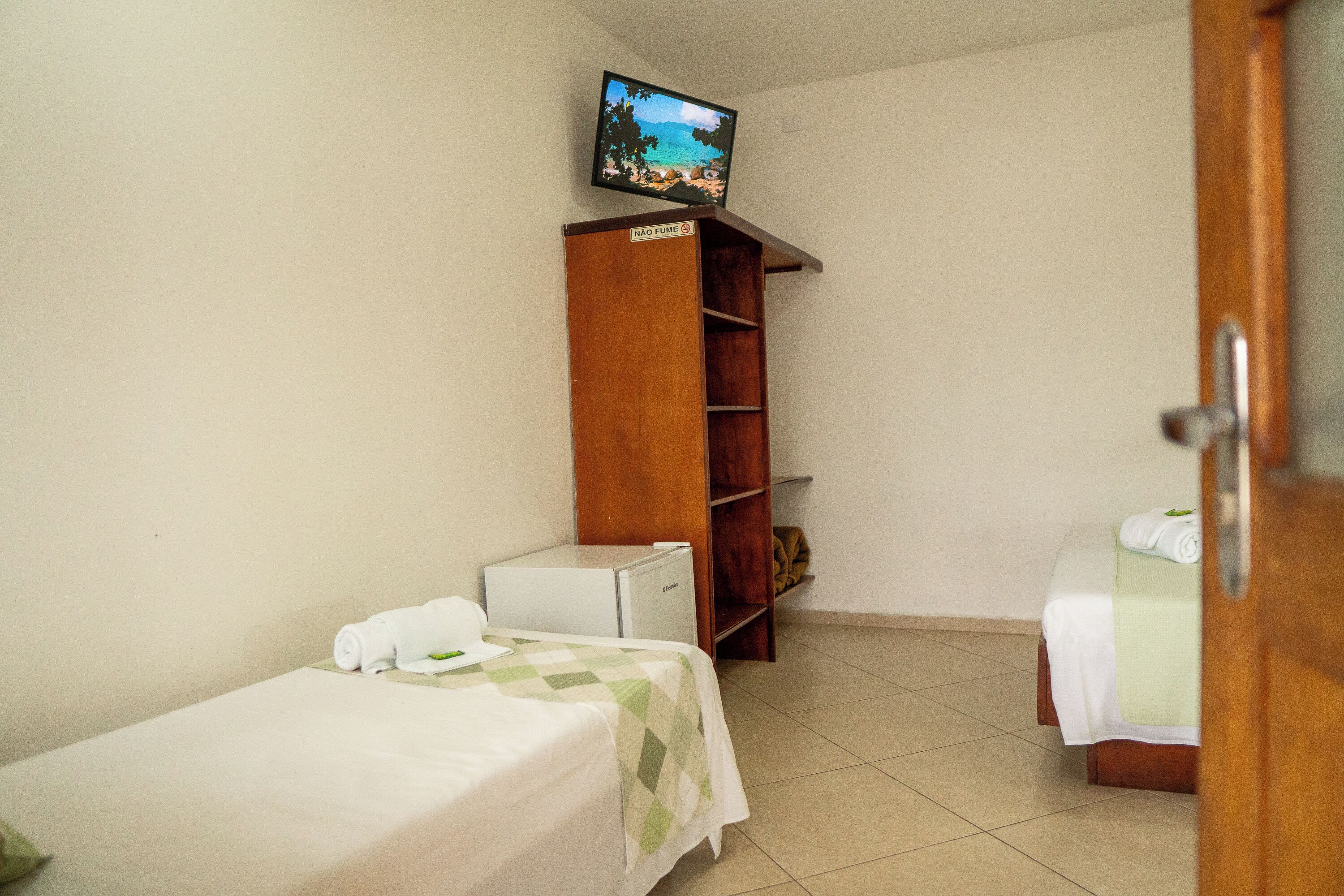 A small hotel room with two single beds, a wooden wardrobe, a television on a shelf, and a small refrigerator.