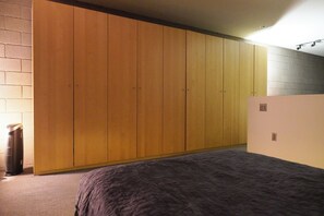 Ein Schlafzimmer mit großem Kleiderschrank, einem Bett mit dunkler Bettdecke und einem Papierkorb.