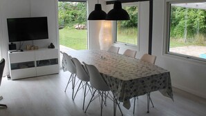 Et moderne spiseområde med et stort bord, hvide stole, et fjernsyn og en bogreol.