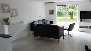 En moderne stue med en sort sofa, et sofabord og et tv-møbel med et fladskærms-tv. Der er billeder i rammer på væggen og en potteplante på skænken.