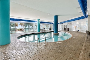 Una piscina interior con suelo de baldosas, pilares azules y vistas a palmeras al exterior.