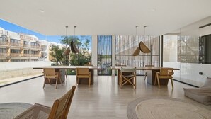 Un espace intérieur vaste et lumineux, avec une longue table et des chaises, de grandes fenêtres et une vue sur les bâtiments environnants.