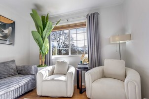 A living room with a grey sofa, two white armchairs, a large plant, a window with curtains, a lamp, and a decorative item on a small table.