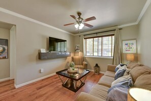 A living room with a ceiling fan, a sofa, a coffee table, a TV mounted on the wall, and a window with curtains. - The Homestead Luxury-cozy 2BR Apartments Near SFA (Nacogdoches)