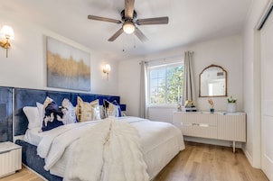 A bedroom with a bed, a ceiling fan, a nightstand, a dresser, a window with curtains, and a wall-mounted mirror.