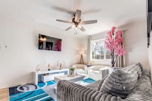 A modern living room with a ceiling fan, a flat-screen TV, a white sofa, a coffee table, and a decorative pink tree.
