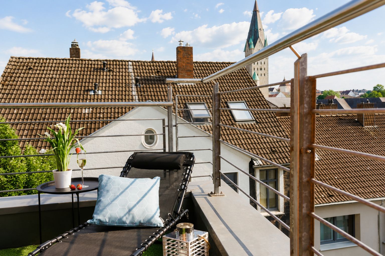 Eine Dachterrasse mit Liegestuhl, einem kleinen Tisch mit Topfpflanze und einem Weinglas, mit Blick auf die Stadtlandschaft.