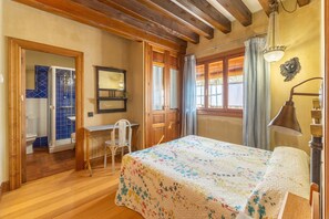 A bedroom with a wooden floor, a bed with a floral bedspread, a desk with a chair, a mirror, and a chandelier.