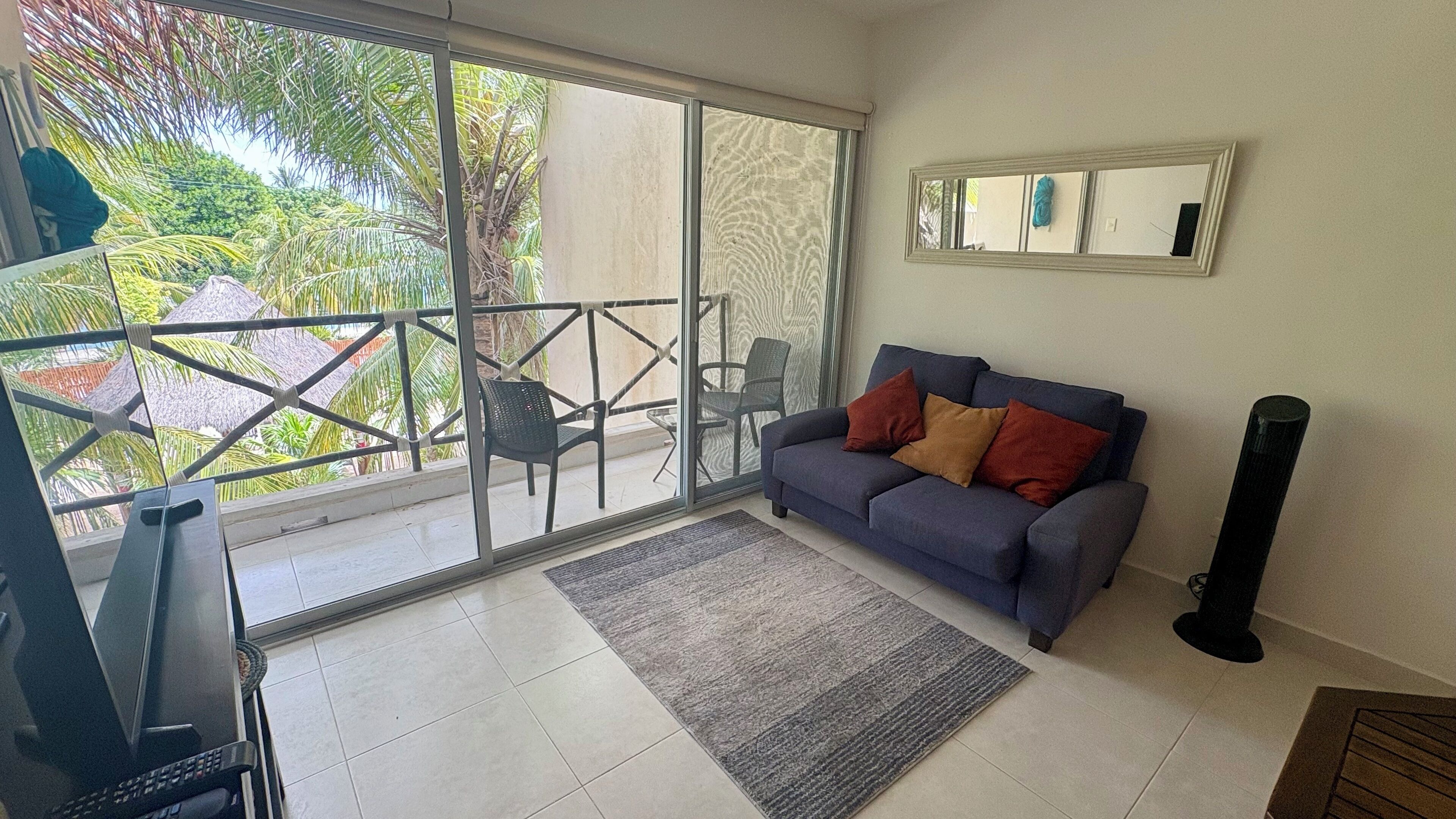 A living room with a grey sofa, a small rug, and a TV. There is a balcony with a glass door and a chair outside.