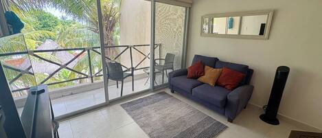 A living room with a grey sofa, a small rug, and a TV. There is a balcony with a glass door and a chair outside.