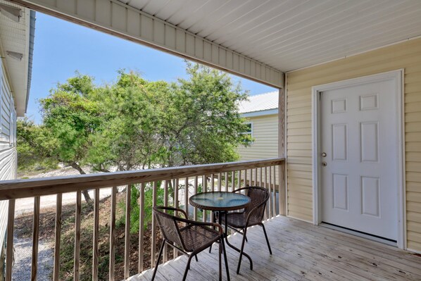 A covered porch with a table and chairs, a white door, and a view of trees and a building in the background. - Whitesands Beach House - North (Gulf Shores)