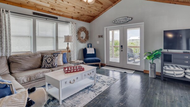 A cozy living room with a wooden ceiling, a sofa, a coffee table, and a TV stand. - Whitesands Beach House - North (Gulf Shores)