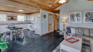 A spacious living area with a wooden ceiling, a dining table with chairs, a sofa, and a coffee table. - Whitesands Beach House - North (Gulf Shores)
