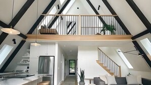 Ein geräumiger Loft mit hoher Decke, freiliegenden Balken, einer Küche mit Kochinsel, einem Essbereich mit einem weißen Kunstfell und einer Treppe, die zu einem erhöhten Wohnbereich führt.