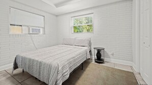 Ein Schlafzimmer mit einem Bett, zwei Fenstern, einem kleinen Tisch und weißen Ziegelwänden.