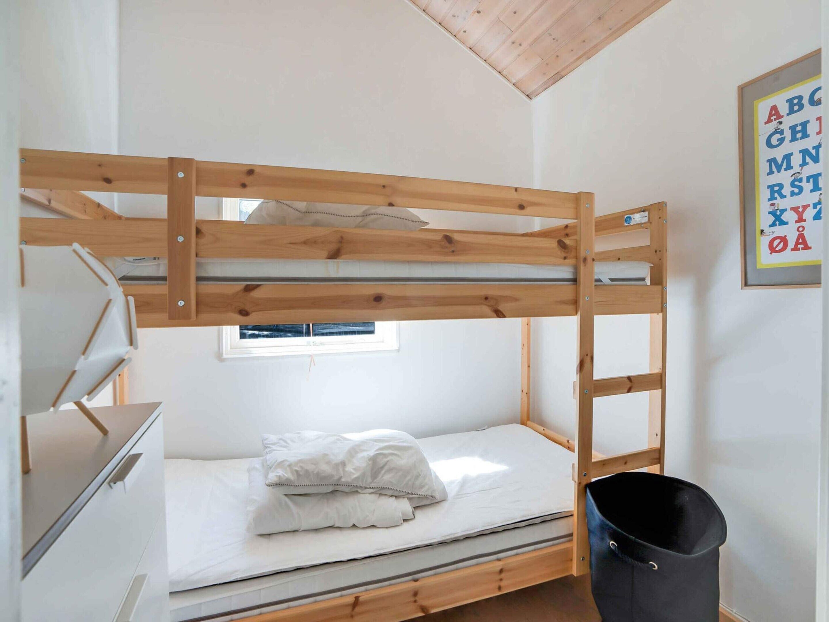 Un letto a castello con struttura in legno, una piccola finestra, una scrivania con cassetti e un cestino nero.