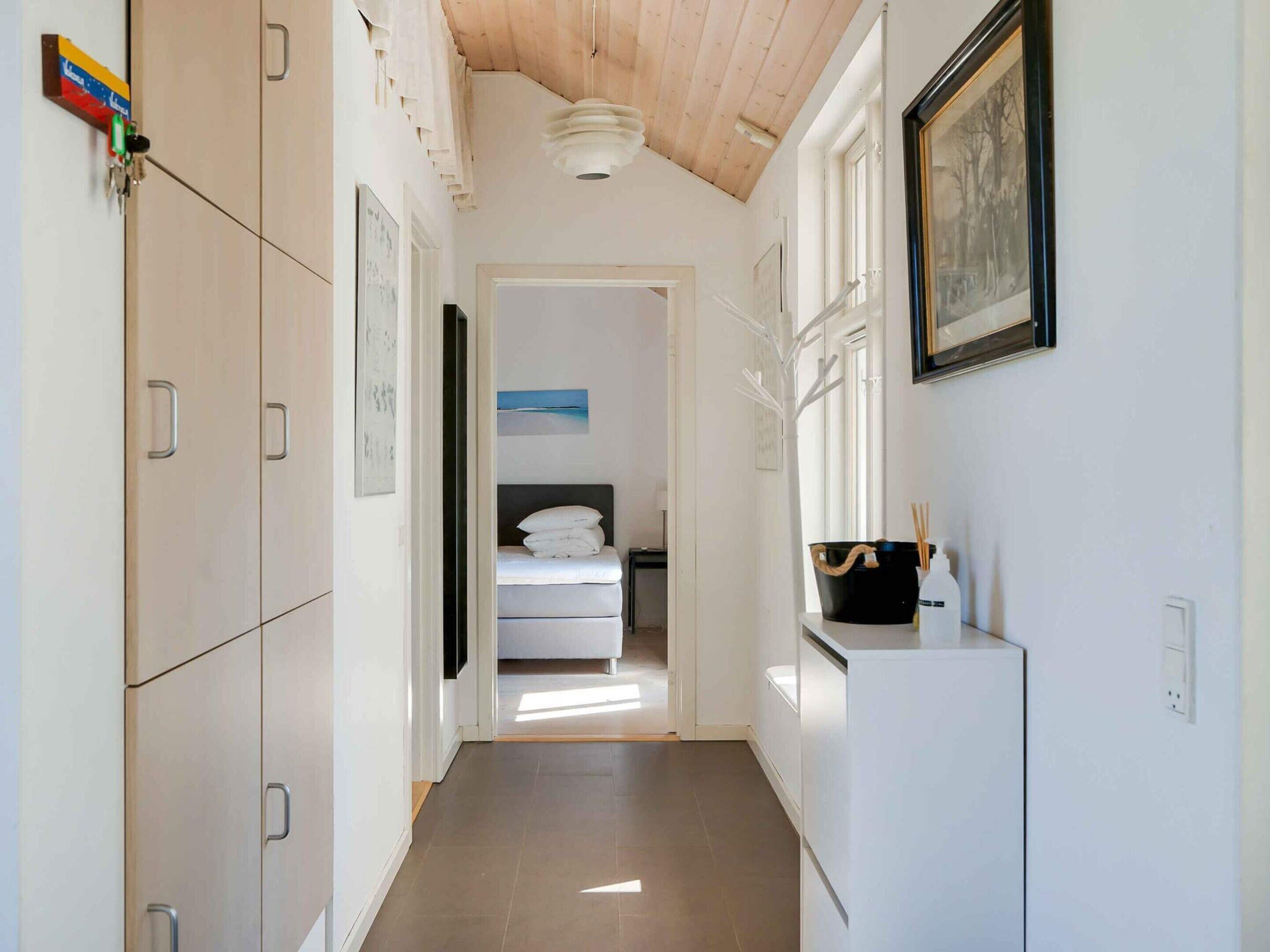 A narrow hallway with a wooden ceiling, a bed with white bedding, and a black bucket with a white bottle on a white cabinet.