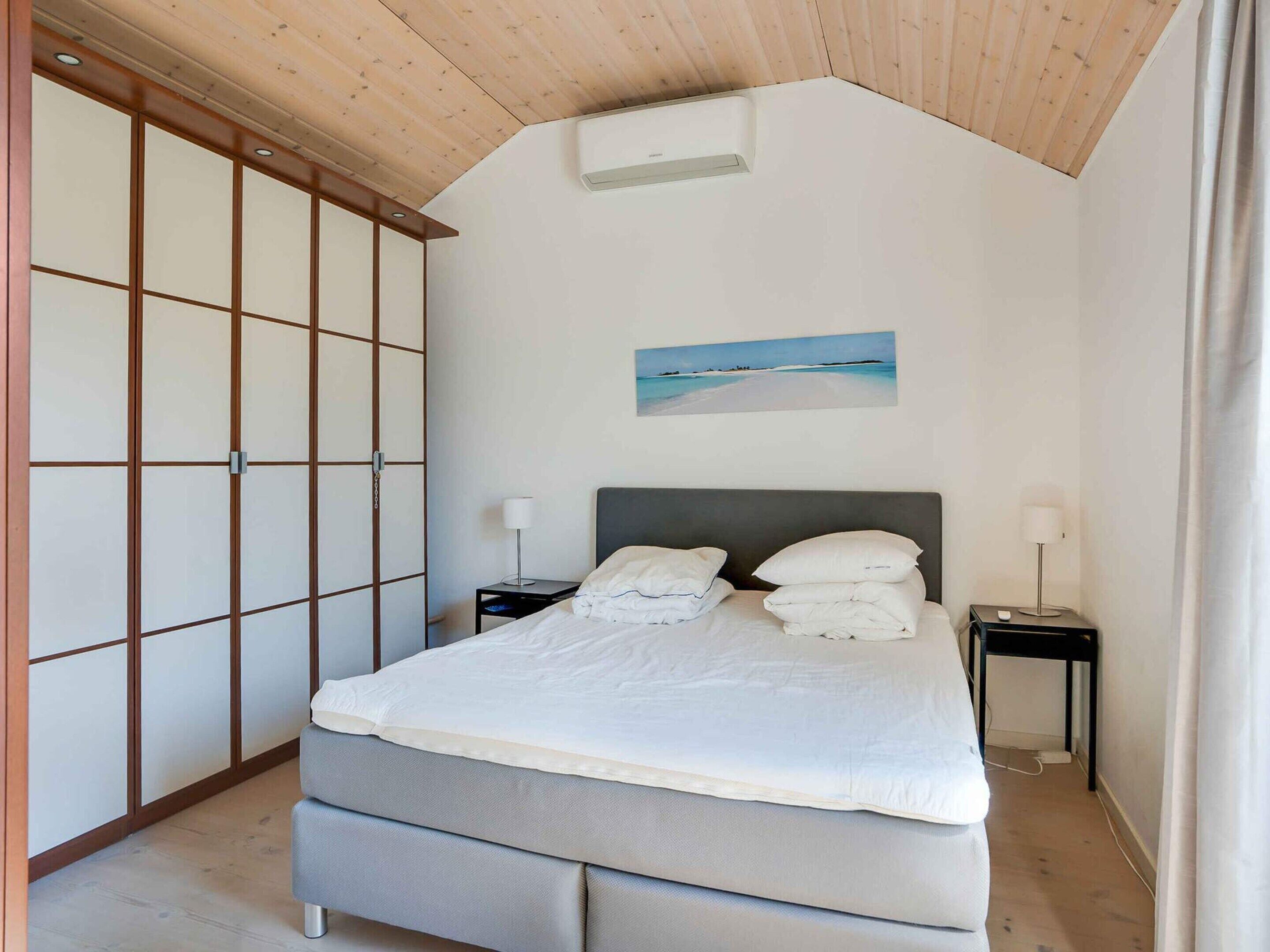 Een slaapkamer met een bed, twee nachtkastjes, een nachtkastje, een wandairconditioner en een schilderij van een strandgezicht.