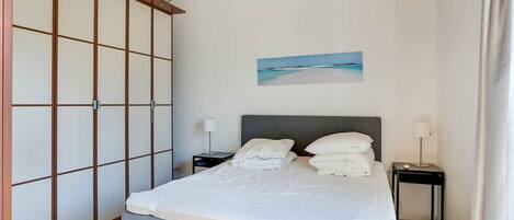 Een slaapkamer met een bed, twee nachtkastjes, een nachtkastje, een wandairconditioner en een schilderij van een strandgezicht.