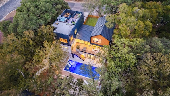Ett modernt hus i två våningar med en takterrass, en swimmingpool och en stor trädgård som omges av träd.
