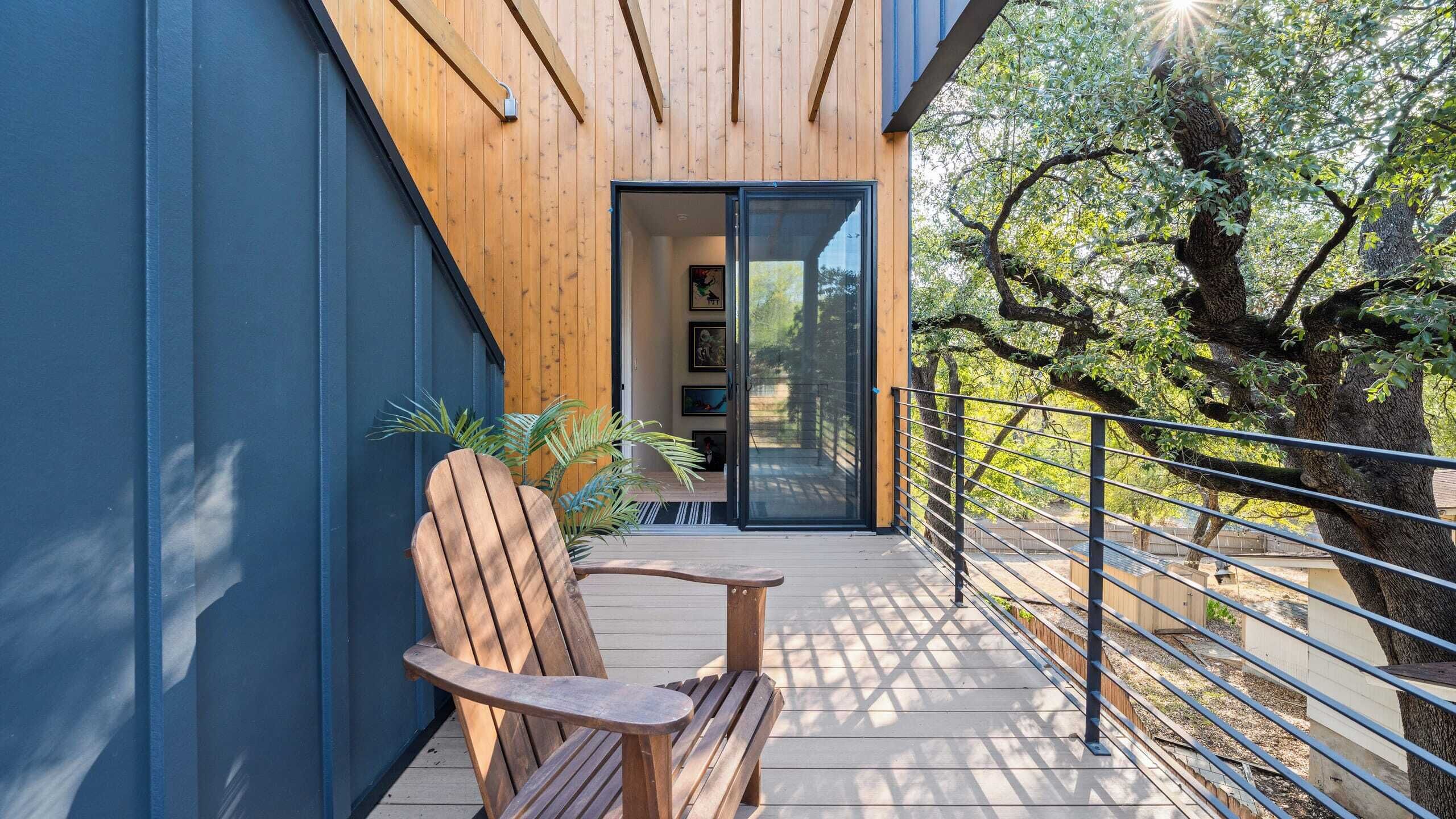 Eine Holzterrasse mit einer Bank und einem Blumentopf, eine Glastür, die zu einem Raum mit gerahmten Bildern führt, und ein Baum im Hintergrund.