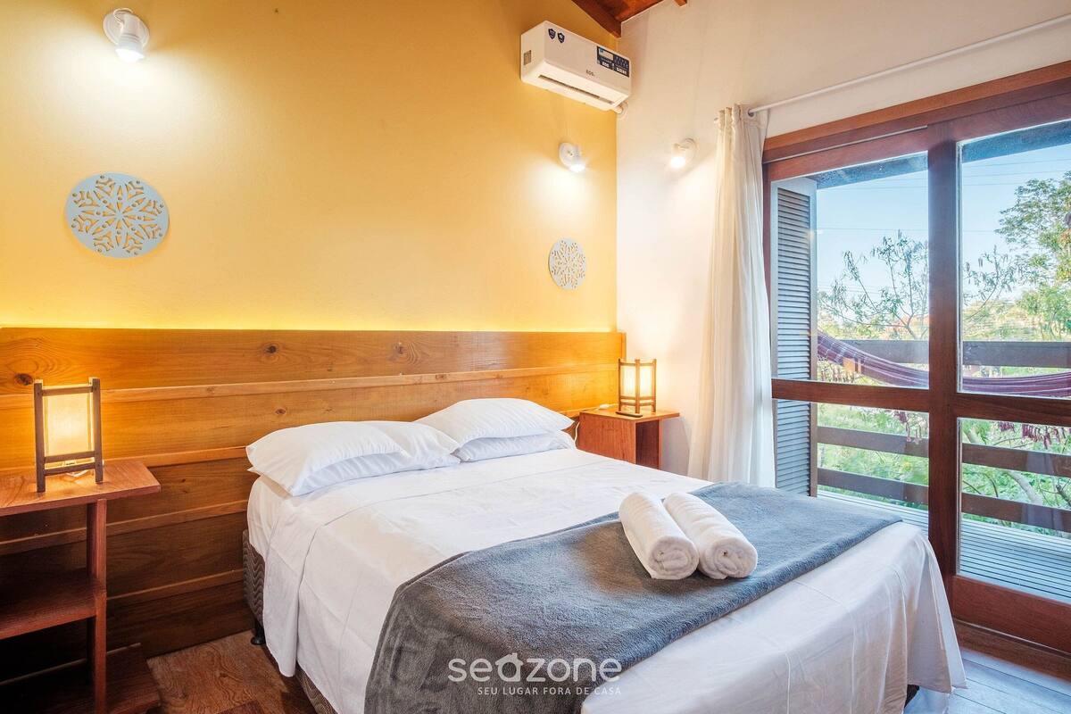 Ein helles Schlafzimmer mit einem Holzkopfende, einem Bett mit weißen Leinen, einem Nachttisch mit Lampe und einem Fenster mit Blick auf Grünflächen.