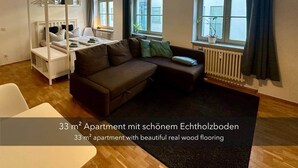En 33 m² lejlighed med trægulv, en seng med himmelseng, sofa, lille bord og store vinduer med gardiner.