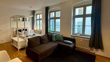 Eine 33 m² große Wohnung mit Holzdielenboden, einem Himmelsbett, einem Sofa, einem kleinen Tisch und großen Fenstern mit Vorhängen.