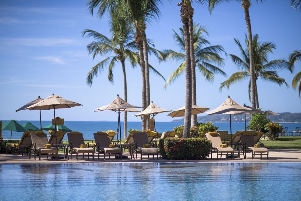 A poolside area with lounge chairs and umbrellas, palm trees, and a view of the ocean.