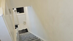 Una escalera alfombrada con barandilla, paredes blancas y un pequeño ventanal en la parte superior.