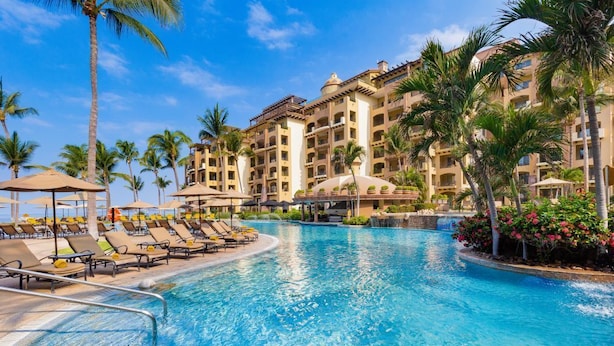 Una piscina de resort con tumbonas, palmeras y un edificio de varios pisos con balcones.