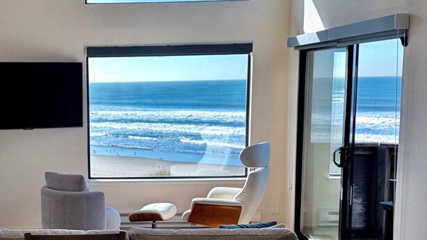 Un salón moderno con una amplia vista al océano, un ventilador de techo y un televisor de pantalla plana montado en la pared.