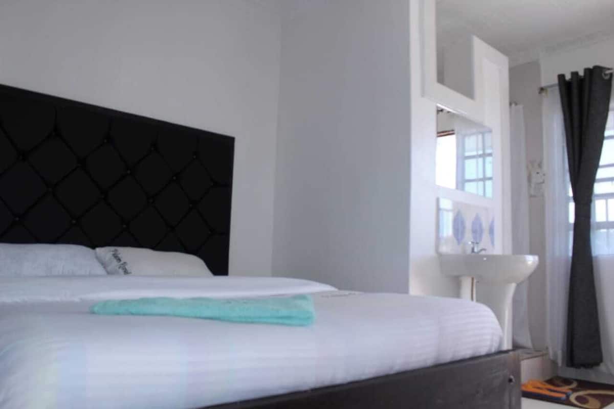 A hotel room with a bed, a white wall, a window with curtains, and a white sink.