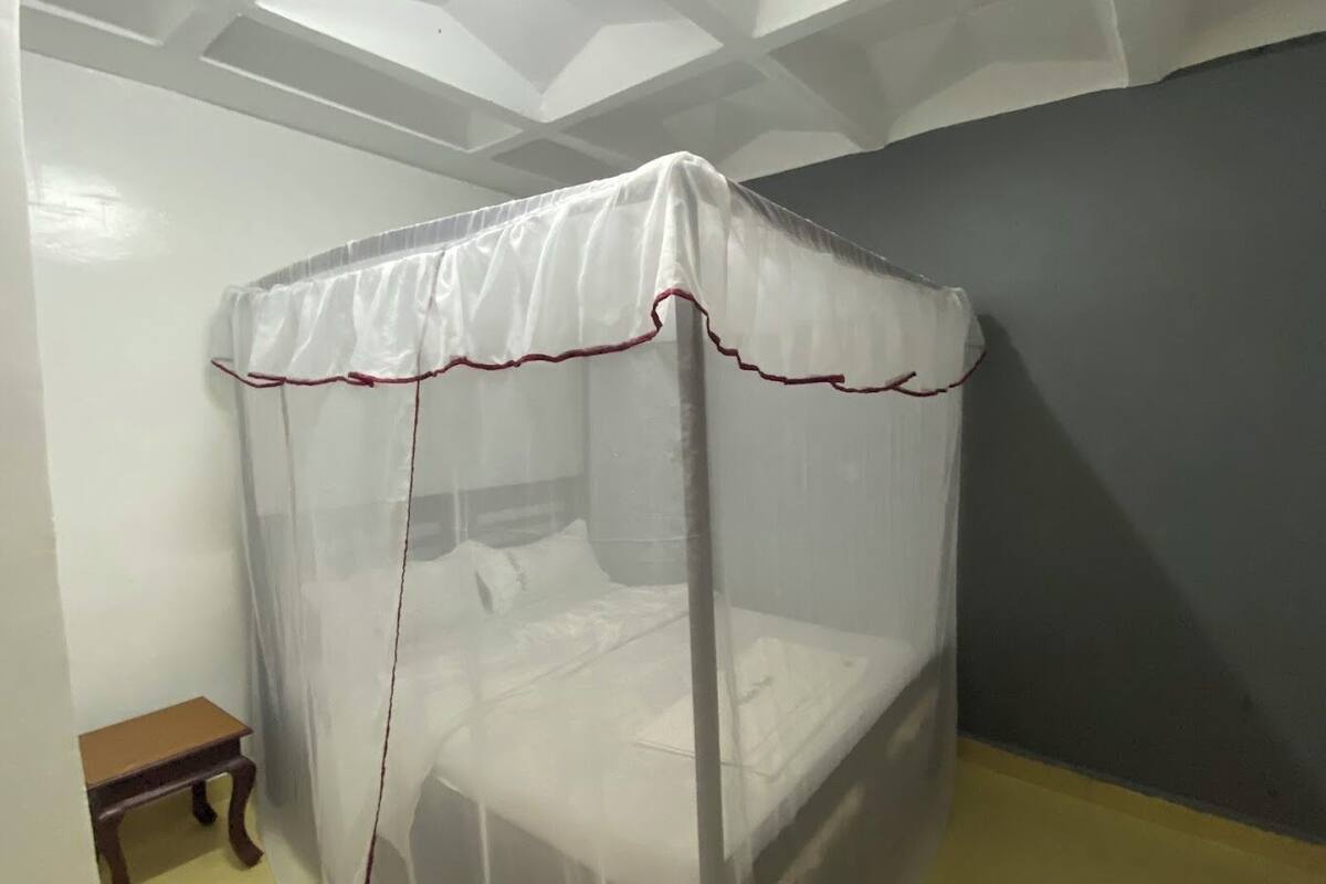 A room with a bed under a mosquito net, a small wooden table, and a plain wall.