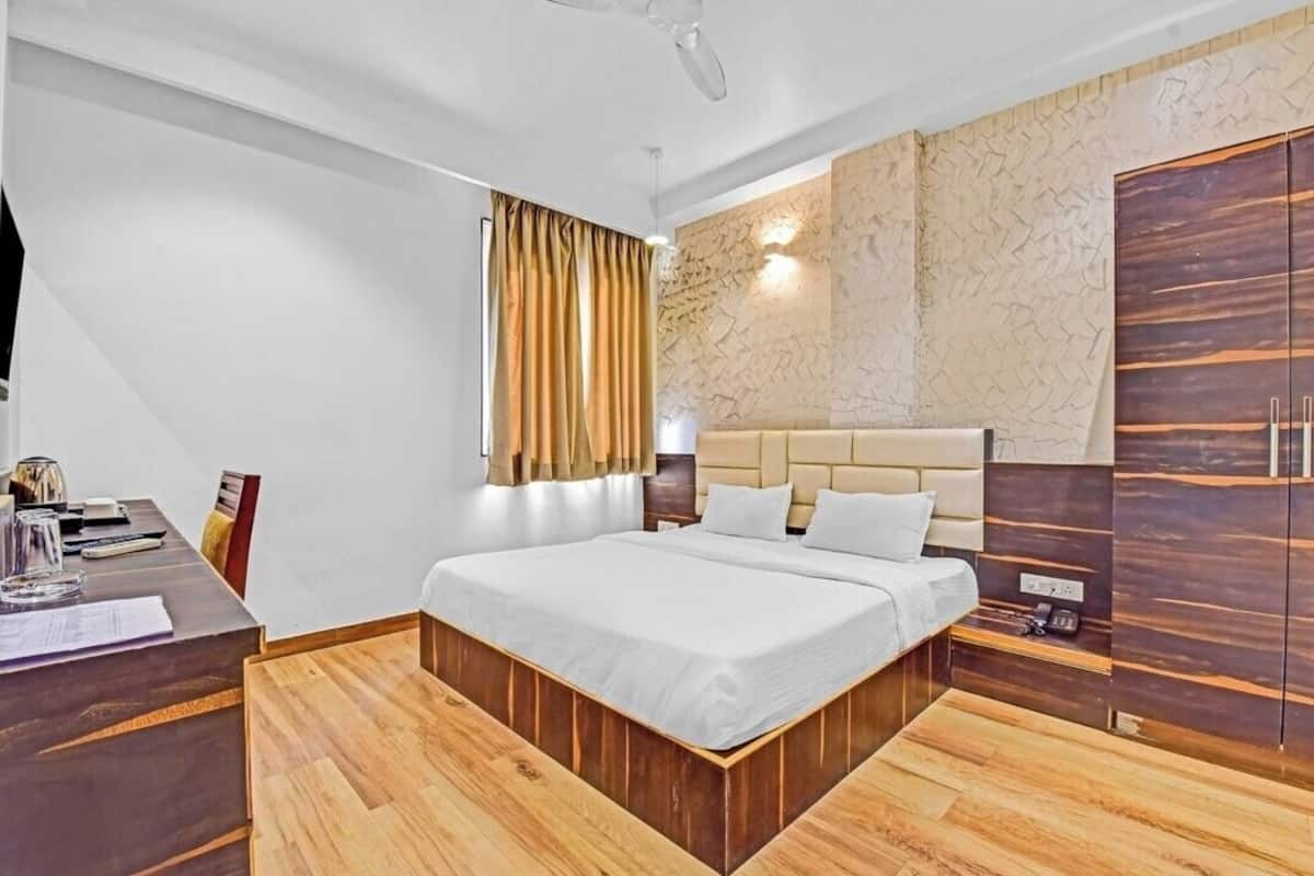 A modern hotel room with a large bed, wooden flooring, a desk with a chair, a wardrobe, and a window with curtains.