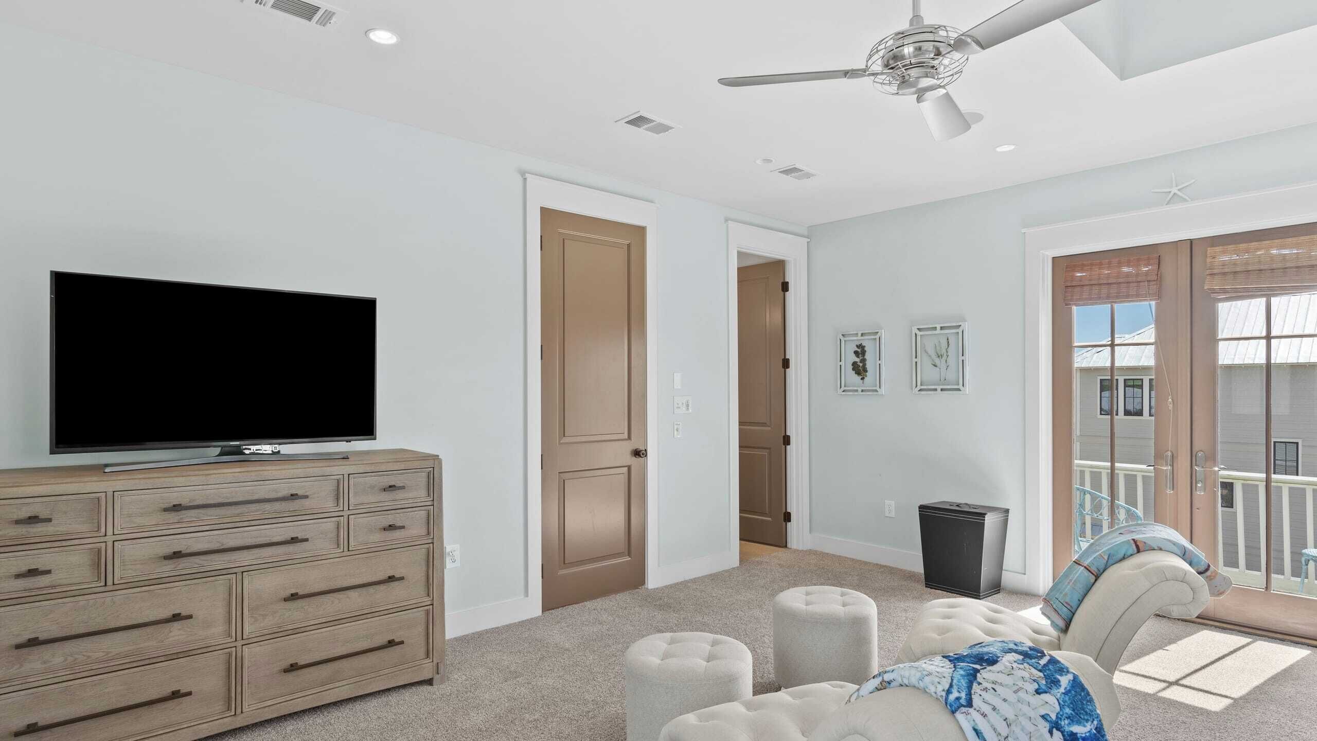 A modern living room with a flat-screen TV, a wooden dresser, a ceiling fan, and a view of the outside balcony.