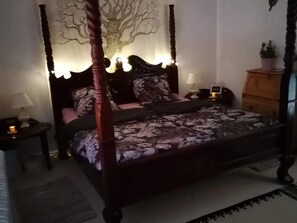A four-poster bed with a floral bedspread, a nightstand with a lamp, a dresser, and a wall-mounted tree decoration.