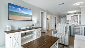 Ein Wohnzimmer mit einem großen Fernseher, einem Holztisch, einem gestreiften Sessel und einer Küchenzeile mit weißen Schränken und einer Steinwandverkleidung.