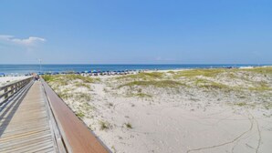 Ein hölzerner Steg führt zu einem Strand mit Dünen und wolkenlosem, blauem Himmel.