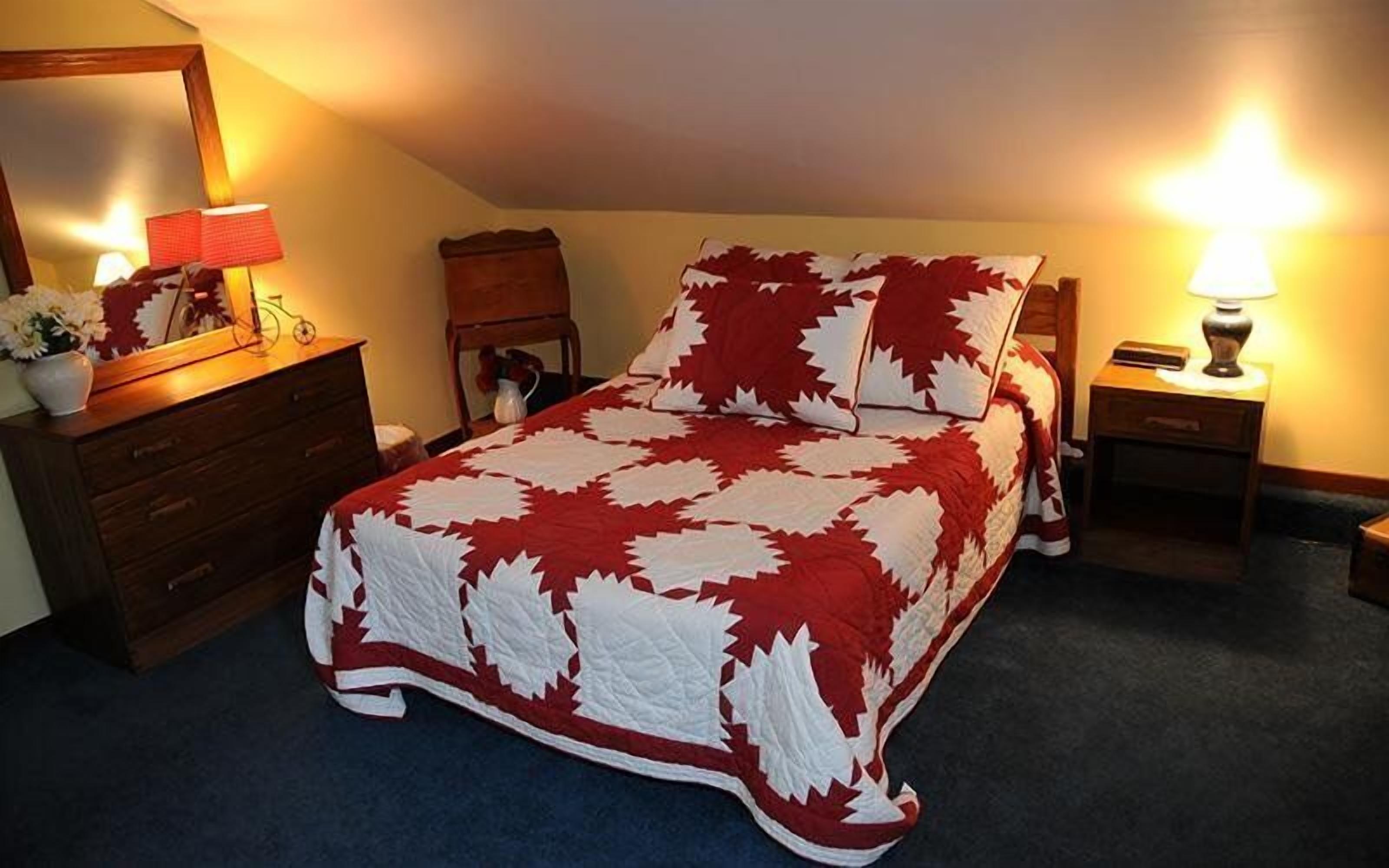 A bedroom with a bed covered in a red and white patterned bedspread, a wooden dresser, a nightstand with a lamp, and a mirror on the wall.