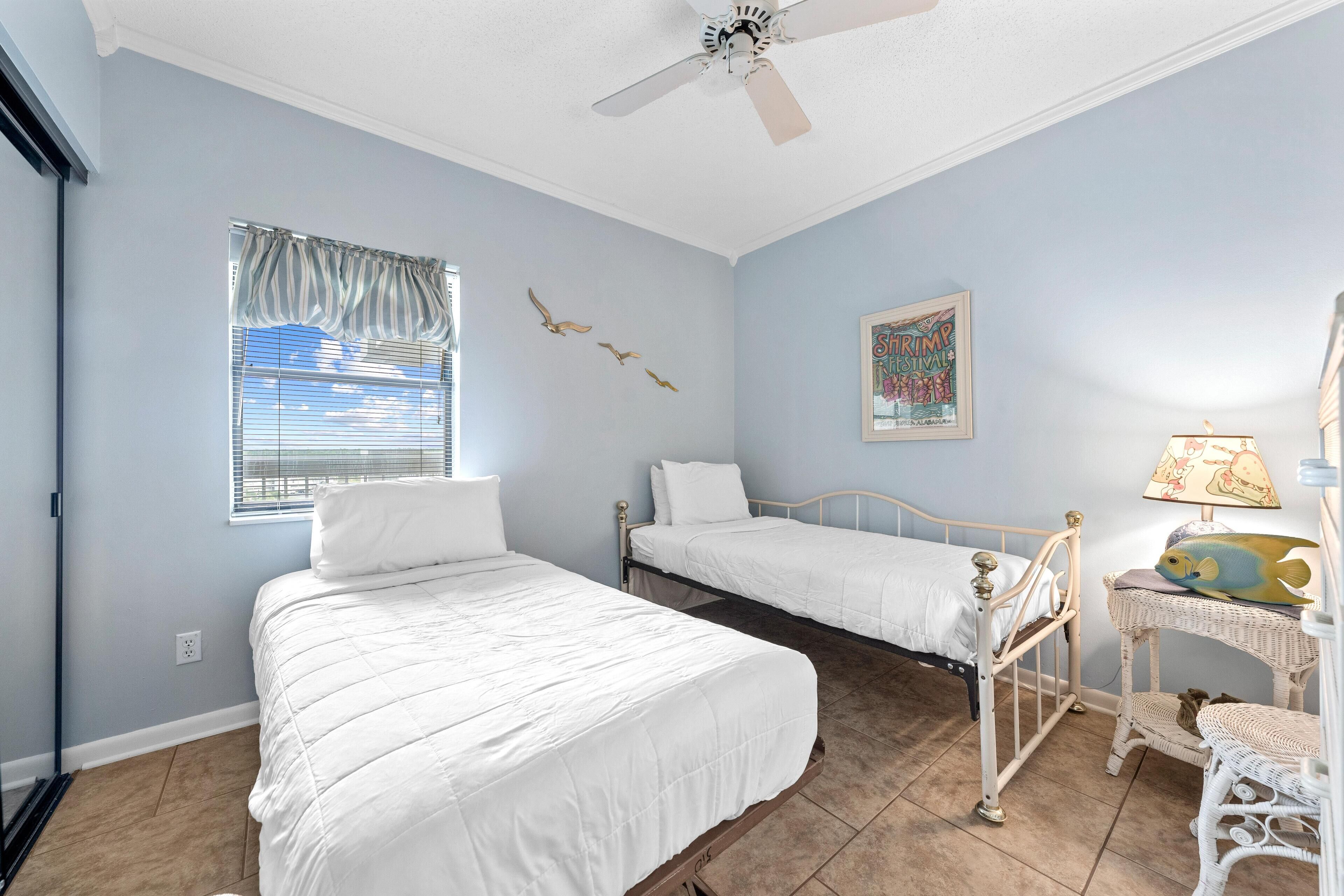 A bedroom with two beds, a ceiling fan, a window with blinds, a wall-mounted artwork, and a wicker side table with a lamp.