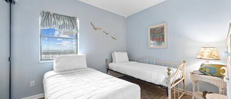A bedroom with two beds, a ceiling fan, a window with blinds, a wall-mounted artwork, and a wicker side table with a lamp.